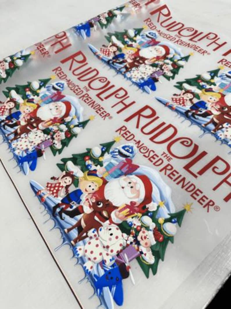 May include: Clear plastic sheet with a repeating pattern of Christmas characters, including Santa Claus, Rudolph the Red-Nosed Reindeer, and elves. The text "Rudolph the Red-Nosed Reindeer" is printed in red. The design features a festive holiday theme.
