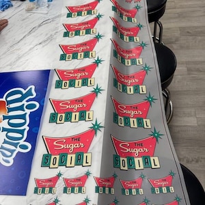May include: Printed sheet featuring repeating retro-style logos for "The Sugar Social." Each logo has a red banner with the text, a teal-colored font, and teal starburst accents. A blue and white Sprite sign is visible.