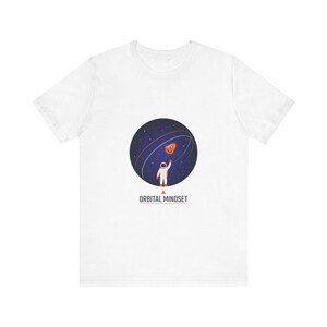 Könnte beinhalten: Weißes T-Shirt mit einer Grafik eines Astronauten, der nach einem Weltraumobjekt in einer sternenübersäten Sphäre greift. Der Text "ORBITAL MINDSET" ist darunter aufgedruckt. Das T-Shirt besteht aus einem weichen, leichten Material.