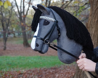 May include: A gray and white hobby horse with a black mane and tail. The horse is wearing a black leather bridle with a silver bit and a beaded browband.