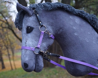 Può includere: Una testa di cavallo di peluche grigia con una briglia viola e nera. Il cavallo ha un occhio nero e un naso nero.