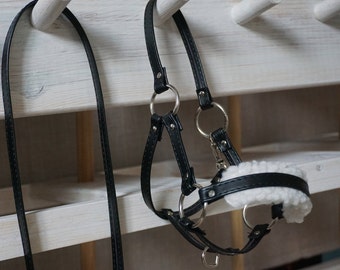 May include: Black leather horse halter with a white fleece noseband hanging on a white wooden rack.