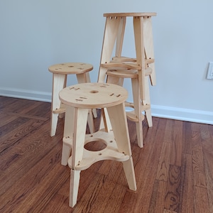 May include: Four wooden stools with a triangular base and a circular seat. The stools are made of light wood and are stacked on top of each other.