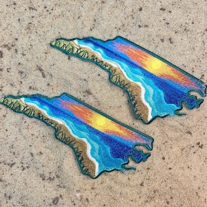 May include: Two embroidered patches in the shape of a coastline, featuring a beach scene with blue water, a yellow sun, and orange and yellow sky. The patches have a green border and are set on a sandy surface.