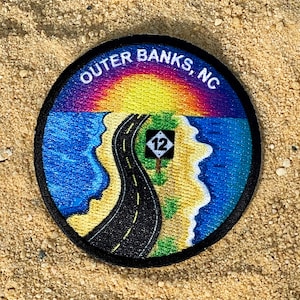May include: A round, black-bordered patch with a colorful image of a road leading to a beach with a sunset in the background. The text "OUTER BANKS, NC" is printed around the top edge of the patch. A diamond-shaped road sign with the number "12" is visible on the road.