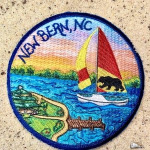 May include: Embroidered patch with a blue border featuring a sailboat on a lake with a bear silhouette on the sail. The patch includes the text "NEW BERN, NC" in blue, with a colorful sunset and landscape background.
