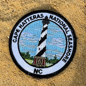 May include: A black and white embroidered patch with a lighthouse, clouds, and the text "Cape Hatteras National Seashore NC".