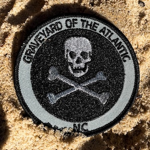 May include: A round black patch with the text "GRAVEYARD OF THE ATLANTIC" and "NC" in white. The patch features a skull and crossbones design in the center, with a gray border. The patch is on a sandy surface.
