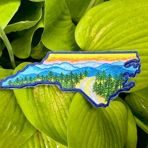 Könnte beinhalten: Gestickter Aufnäher in Form von North Carolina, mit einem Landschaftsdesign mit Bergen, Bäumen und einer kurvenreichen Straße. Der Aufnäher hat einen blauen Rand und ist vor einem Hintergrund aus grünen Blättern platziert.