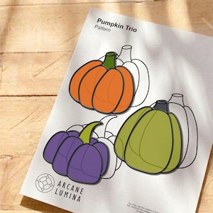 May include: A printed paper sheet titled Pumpkin Trio Pattern featuring three illustrated pumpkins in orange, purple, and olive green with black outlines. The design includes line art sketches of pumpkins and the text Arcane Lumina at the bottom.