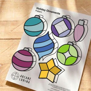 Op de afbeelding: Een geprint patroonblad getiteld Holiday Ornaments van Arcane Lumina met diverse ontwerpen in glas-in-loodstijl, waaronder een turquoise bol, paarse ruit, roze peervorm, blauwe druppel, groene cirkel en een gele ster met zwarte omlijningen.