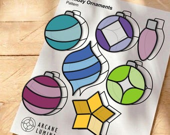 Könnte beinhalten: Ein gedrucktes Vorlagenblatt mit dem Titel Holiday Ornaments von Arcane Lumina, das verschiedene Designs im Buntglasstil zeigt, darunter eine türkisfarbene Kugel, eine violette Raute, eine rosa Birne, einen blauen Tropfen, einen grünen Kreis und einen gelben Stern mit schwarzen Umrissen.