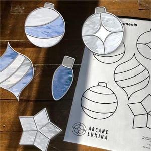 May include: A selection of stained glass Christmas decorations in diverse shapes, such as a star, teardrop, and bulb. The ornaments have silver edging and blue and white glass. A sheet with ornament outlines and the text "Arcane Lumina" is also visible.
