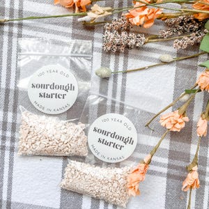 Pode incluir: Duas pequenas sacolas brancas com etiquetas brancas que dizem "100 year old sourdough starter made in Kansas". As sacolas contêm pequenos pedaços de fermento branco. As sacolas estão sobre uma superfície xadrez cinza e branca com flores secas.