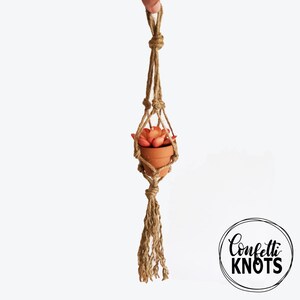 May include: A small terracotta pot with a succulent plant inside is hanging from a macrame plant hanger made of natural jute rope. The hanger has a tassel at the bottom. The text "Confetti Knots" is visible in the lower right corner of the image.