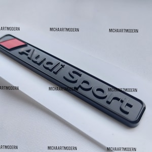 May include: Black and red Audi Sport emblem with a raised, textured design. The emblem is rectangular and has a red stripe at the top.