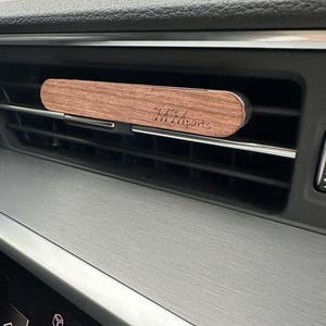 May include: A wooden car air freshener with the text "M&M Parts" engraved on it. The air freshener is attached to a car's air vent.