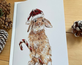 May include: A brown rabbit wearing a red Santa hat and holding a candy cane. The rabbit is sitting on a white background with a snowy texture.
