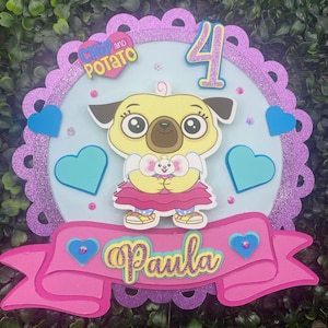 Könnte beinhalten: Ein rosa und lilafarbener Tortendekor mit den Figuren Chip und Potato. Der Dekor hat einen blauen Kreis mit einem gelben Hund, der eine weiße Maus hält. Die Zahl 4 befindet sich in der oberen rechten Ecke des Kreises. Der Text "Chip and Potato" befindet sich in der oberen linken Ecke des Kreises. Ein rosa Banner mit dem Text "Paula" befindet sich am unteren Rand des Dekors.