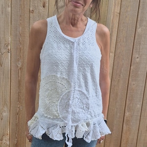 May include: White tank top with eyelet and lace detailing. The top has a round neckline, a drawstring, and a ruffled hem. The design incorporates circular lace elements, creating a layered, textured look. The top is paired with blue jeans.