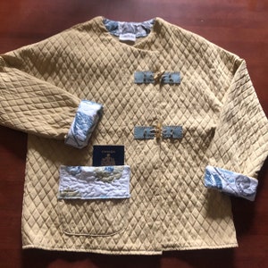 Pode incluir: Uma jaqueta acolchoada de cor amarela clara com um padrão de losangos. A jaqueta tem um forro floral azul e branco e duas grandes fivelas douradas na frente. A jaqueta tem um bolso no lado esquerdo com um passaporte azul dentro.