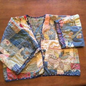 May include: A patchwork quilt jacket featuring a floral design. The jacket is made from a variety of fabrics in shades of blue, pink, yellow and white. The sleeves are long and have a blue and white trim. The jacket is quilted with a floral pattern.