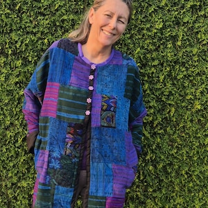 May include: A long-sleeved jacket featuring a patchwork design in shades of blue, purple, and dark green. The garment has a round neckline, a front button closure with small light pink flower-shaped buttons, and a solid purple inner lining.