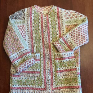 May include: A long-sleeve quilted jacket featuring a patchwork design with various floral and geometric patterns in shades of pink, green, and cream. The jacket has a round neckline, a front button closure with metallic buttons, and two large patch pockets.