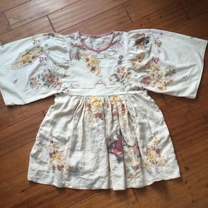 May include: Cream-coloured dress with floral embroidery. The dress has wide sleeves and a gathered skirt. The embroidery includes pink, yellow, and brown flowers. The neckline is trimmed with pink fabric.
