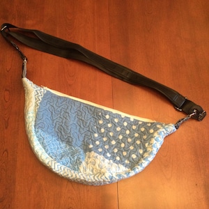 May include: A blue and white quilted fabric half-moon shaped bag with a black adjustable strap.