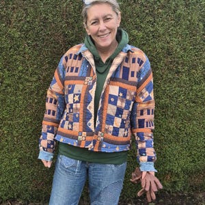Peut inclure: Veste matelassée avec un motif patchwork bleu, orange et beige. La veste a une fermeture boutonnée et un col. La personne porte un sweat à capuche vert, un jean bleu et des gants marron.
