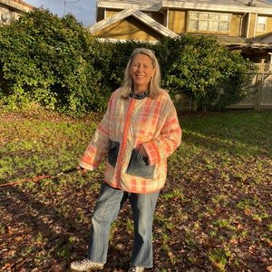 May include: A woman wearing a pink and white checked quilted jacket with a denim patch pocket. She is standing in a grassy area with fallen leaves.