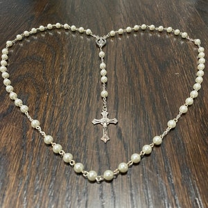 Puede incluir: Un rosario de perlas blancas con un colgante de cruz de plata.