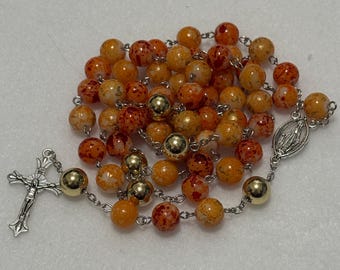 May include: A rosary featuring orange and red speckled beads, interspersed with smooth gold-tone metal beads. The chain is silver-tone metal, connecting to a silver-tone crucifix and a small oval medal depicting a figure on one side.