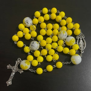 May include: A yellow rosary with white beads featuring black accents. The rosary has a silver crucifix and a silver charm of the Virgin Mary.