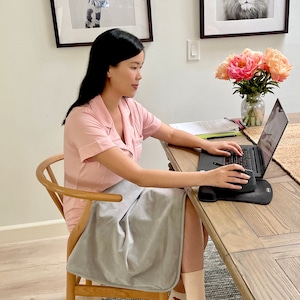 Peut inclure: Une femme vêtue d'un haut de pyjama rose est assise à un bureau en bois avec un ordinateur portable et une couverture grise drapé sur ses jambes. Elle utilise une souris sans fil pour naviguer sur l'ordinateur portable.