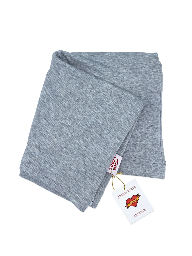 May include: A gray, folded, soft, cotton scarf with a tag that says "The Cozy Warrior" and has a red heart with the word "Warrior" inside. The tag also says "Wear it proudly, be a cozy rebel, but it doesn't have to be a lonely one."