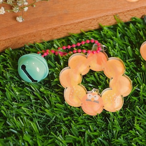 Op de afbeelding: Een teal bel charm met een roze kralenketting bevestigd aan een transparante oranje en gele donut charm met een cartoon gezicht en de tekst "2017" erop.