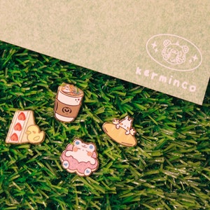 Peut inclure: Quatre épingles en émail représentant des personnages de dessins animés sur un fond vert. Les épingles représentent une tasse de café, une part de gâteau, un nuage avec un personnage à l'intérieur et un personnage chevauchant une baguette.