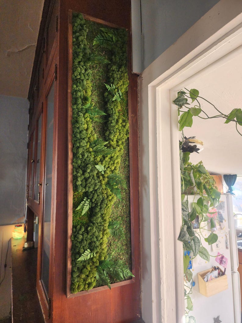 May include: A framed vertical moss wall art piece with a variety of green moss and ferns. The piece is mounted on a wooden wall.