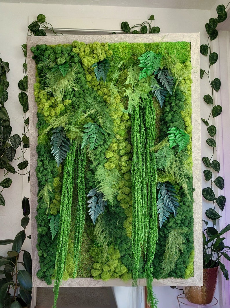 May include: A framed moss wall art piece featuring a variety of green moss and ferns. The moss is arranged in a natural, organic pattern, creating a lush and textured look. The frame is a light wood colour.