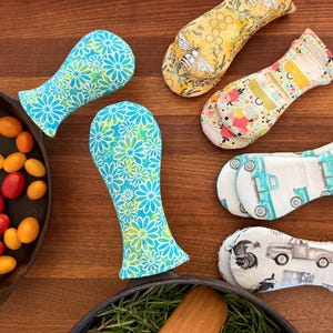 May include: A collection of colorful, patterned pot handle covers. Designs include bees on yellow, floral patterns, and trucks. The covers are designed to protect hands from hot cookware. The covers are made of fabric.