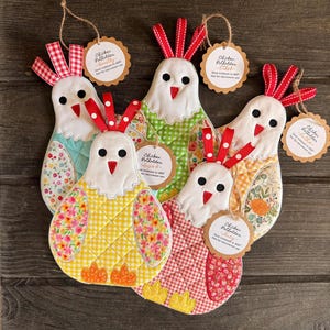 May include: A collection of five chicken-shaped pot holders in various colors and patterns. Each pot holder features a white head with a red beak and a red ribbon comb. The pot holders have a quilted design and are adorned with tags.