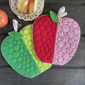 May include: Three apple-shaped quilted pot holders in red, green, and floral patterns. Each has a green leaf and a loop for hanging. A whole apple and a bowl of apple slices are also in the image, suggesting a kitchen theme.