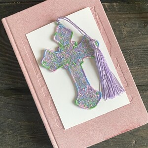 May include: A pastel-colored embroidered cross bookmark with a lavender tassel rests on a pink book with a white card. The cross features intricate floral patterns in shades of green, blue, and purple. The bookmark is a decorative accessory for books.