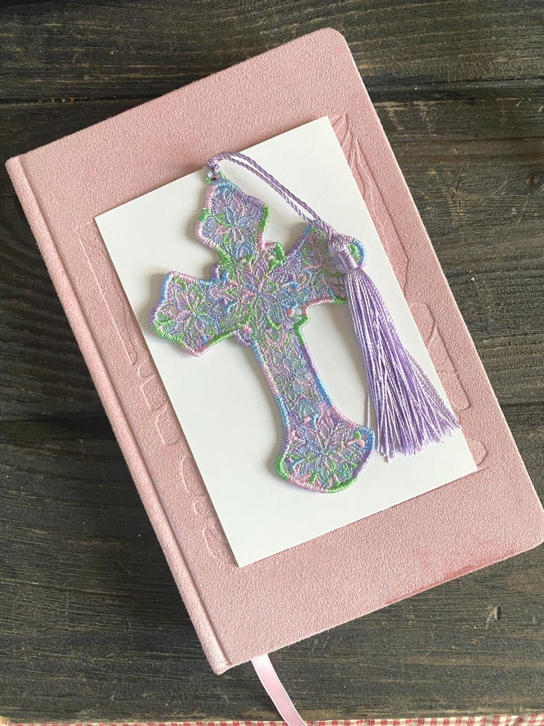 May include: A pastel-colored embroidered cross bookmark with a lavender tassel rests on a pink book with a white card. The cross features intricate floral patterns in shades of green, blue, and purple. The bookmark is a decorative accessory for books.
