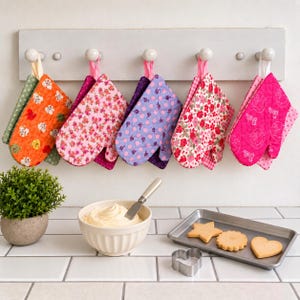 Puede incluir: Cinco manoplas de cocina acolchadas con varios estampados florales y de lunares cuelgan de un perchero de madera. Debajo, un cuenco blanco contiene crema, una espátula de metal y una bandeja metálica con tres galletas y un cortador.