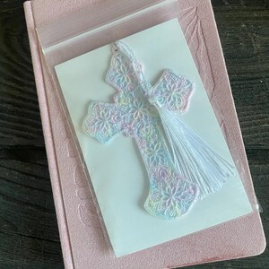 May include: A pastel-colored, embroidered cross bookmark with a white tassel, packaged on a white card. The bookmark is placed on a pink journal with a matching ribbon.