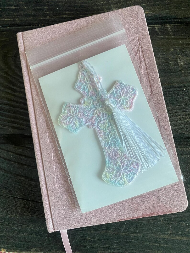 May include: A pastel-colored, embroidered cross bookmark with a white tassel, packaged on a white card. The bookmark is placed on a pink journal with a matching ribbon.