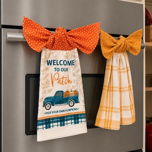 Può includere: Due strofinacci da cucina appesi con la parte superiore a forma di fiocco. Lo strofinaccio a sinistra è color crema con un camion blu che trasporta zucche, il testo "WELCOME TO OUR Patch PICK YOUR OWN PUMPKINS" e un fiocco arancione a pois.